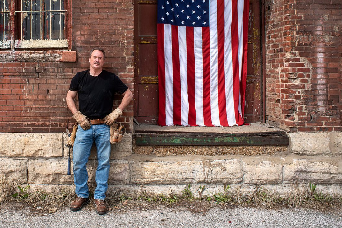 Veteran construction worker leaning on brick wall with American flag