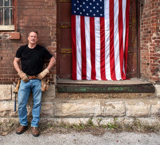 Veteran construction worker leaning on brick wall with American flag